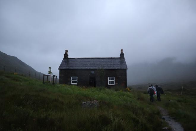 Stone bothy with two chimneys emerging out of the fog, two people walking up to it