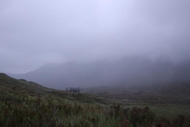 A lone bothy barely visible through thick highland mist at the bottom of a glen