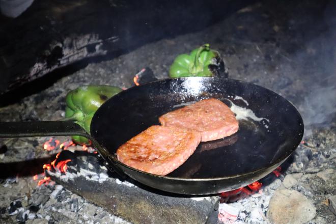 Sausages frying in a pan next to green peppers, balanced over the campfire